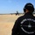 A member of the Pacific Air Forces Demonstration Team stands on a tarmac wearing a black uniform jacket and cap labeled 'F-16 DEMO', with hearing protection headset. In the background, two F-16 fighter jets are parked on the flight line with ground crew personnel nearby. A vapor trail or smoke streak is faintly visible in the blue sky above.