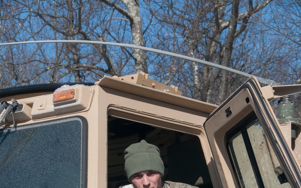 A U.S. Army soldier wearing ACU (Army Combat Uniform) pattern camouflage and an olive drab beanie is seated in or exiting the cab of a tan military tactical truck. The soldier's name tape reads 'RINGE' and the 'U.S. ARMY' tape is clearly visible on the uniform. The vehicle is marked with the identifier 'HZ 113' and appears to be a LMTV or similar military cargo/utility truck.