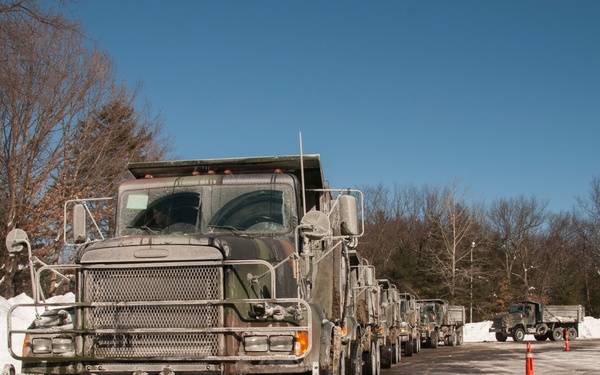 A convoy of military camouflage-painted dump trucks is lined up in a snow-covered parking area or staging ground. The vehicles appear to be undergoing washing or decontamination, evidenced by water and soap residue visible on the lead truck. The scene suggests a winter military operation or post-mission vehicle maintenance activity.