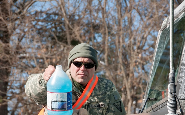 A U.S. Army soldier in camouflage uniform and olive green beanie is holding a gallon jug of blue windshield washer fluid while standing in or beside a military vehicle. The vehicle appears to be a military truck or HMMWV (Humvee) with snow and ice residue on its hood and windshield. The scene takes place outdoors in winter conditions with bare trees visible in the background.