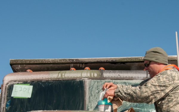 Two military personnel in camouflage uniforms are working on or around a heavily mud-covered Freightliner military truck. One soldier is standing on the front of the vehicle appearing to clean or work on the windshield, while another soldier is visible through or beside the windshield. The scene takes place outdoors under a clear blue sky.