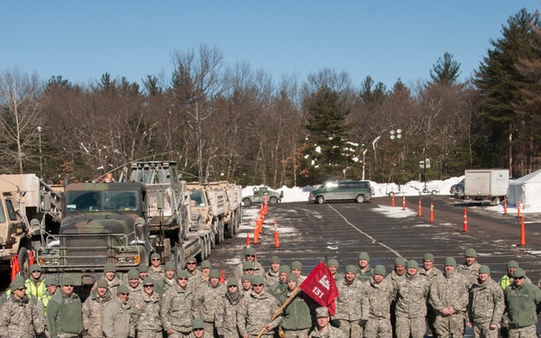 A large group of military personnel in camouflage uniforms pose for a group photograph in a parking lot during winter conditions. They are assembled in front of several military vehicles including heavy transport trucks, with snow visible in the background. A soldier in the front center holds a red unit guidon flag bearing the number '151'.