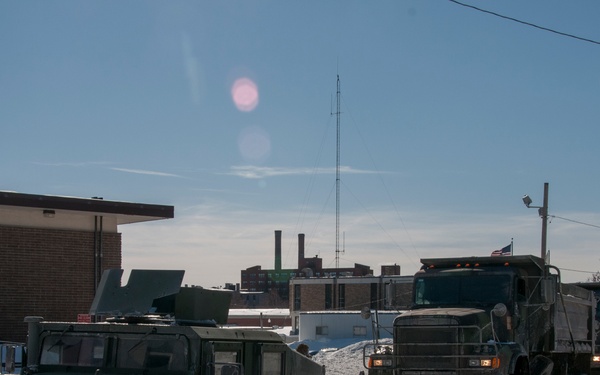 A convoy of military vehicles is traveling along a snow-covered road in what appears to be a small urban area during winter. The convoy consists of at least one HMMWV (Humvee) in the foreground and two larger military trucks behind it. A banner on a nearby pole references 'Northern Essex Community College - Lawrence Campus,' suggesting this is Lawrence, Massachusetts.