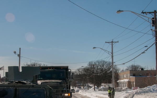 Military vehicles, including a Humvee and a large military dump truck, are operating on a snow-covered urban road following a significant snowstorm. Large snow banks line both sides of the road, and a person in a high-visibility vest is visible near the roadside. An American flag is visible in the background near what appears to be a government or municipal building.
