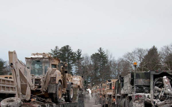 A convoy of military vehicles is staged in a snow-covered parking area or staging ground during winter conditions. The vehicles, including flatbed trailers carrying heavy construction equipment such as a front-end loader, appear heavily coated in mud and road grime. The scene suggests a military unit returning from or preparing for a field operation in cold-weather conditions.