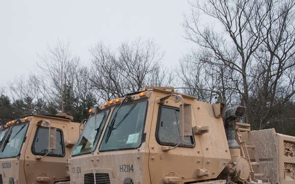 Multiple tan/desert-camouflage military trucks, identified as LMTV/FMTV (Family of Medium Tactical Vehicles), are parked in a lot during overcast winter conditions. The foremost vehicle bears the marking 'HZ114' and unit designation '131 EN', with a dump truck bed configuration visible. The vehicles appear to be staged, possibly for a snow removal or emergency response operation given the wintry environment.