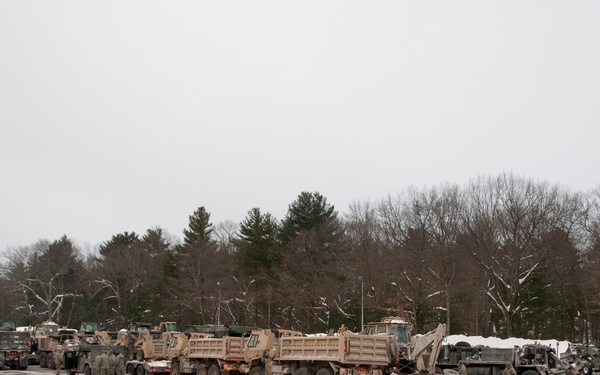 A convoy of military vehicles is staged in a large paved parking area during winter conditions, with snow visible on the ground and surrounding landscape. Multiple tan/olive-drab military trucks, including what appear to be HEMTT (Heavy Expanded Mobility Tactical Truck) dump variants and other heavy equipment, are lined up in formation. Several uniformed personnel are visible near the vehicles, suggesting a pre-mission staging or equipment inspection.
