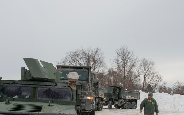 A military convoy of olive-drab vehicles is staged in a snow-covered area during winter conditions. A soldier in camouflage uniform and a green jacket walks alongside the vehicles, which include a Humvee in the foreground and at least two larger military trucks behind it. The scene appears to be a National Guard winter operations or emergency response deployment.