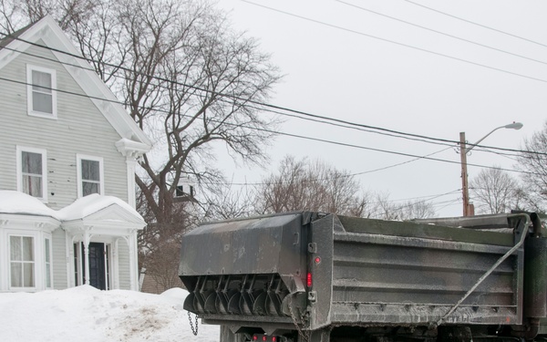 A military-style dump truck is operating on a residential street during or after a significant snowstorm. A person in military/utility fatigues and a knit cap is directing or signaling the truck near a large snow pile. The scene appears to be a snow removal or emergency response operation in a New England-style residential neighborhood.