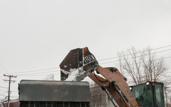 A CASE wheel loader is actively dumping a bucket of snow into the back of a large dump truck during winter snow removal operations on a public road. The scene depicts post-snowstorm cleanup in what appears to be a small town or suburban area, with significant snow accumulation visible along the roadside. Overcast skies, bare trees, and snow-covered surroundings confirm winter conditions.