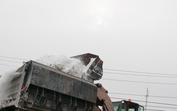 A CASE front-end loader is actively dumping a bucket of snow into the bed of a large dump truck on a road during winter snow removal operations. The scene appears to be in a small town, with a Memorial Building and a red residential structure visible in the background. Significant snow accumulation is visible along the roadside, indicating recent heavy snowfall.