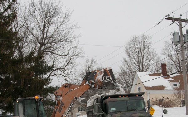 A CASE wheel loader is actively loading snow into a military-style camouflage dump truck during winter snow removal operations at a residential street intersection. A person in military-style camouflage clothing stands to the right, observing the operation. Significant snow accumulation is visible along the roadside, indicating a major snowstorm cleanup effort.