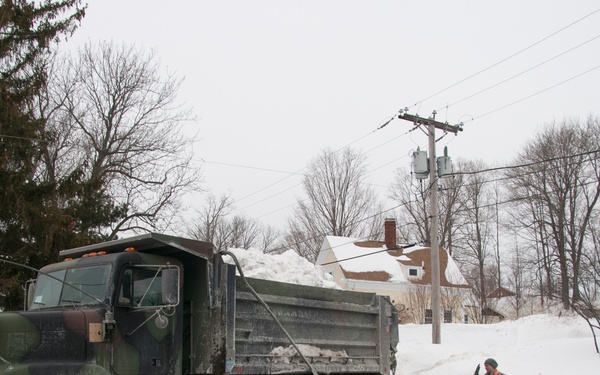 A military dump truck loaded with snow is being directed by a soldier in camouflage uniform on a residential street following a significant snowstorm. The truck appears to be part of a snow removal operation, with large snow piles visible along the roadside and in yards. The scene depicts a military-assisted civilian snow emergency response in what appears to be a northeastern U.S. suburban neighborhood.