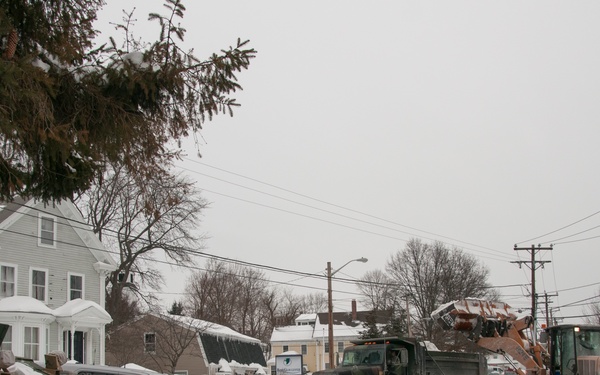 A winter snow removal operation is underway on a residential street with heavy accumulation of snow. Military-style dump trucks and a CASE front-end loader are actively loading and hauling snow, while a police officer and police SUV are present, likely directing traffic or providing oversight. The scene suggests a significant post-blizzard municipal snow removal effort in a northeastern US residential neighborhood.