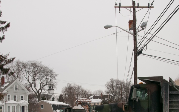 A winter snow removal operation is underway on a residential street, featuring military-style dump trucks in camouflage paint scheme clearing snow after a significant snowstorm. Multiple vehicles are staged along the road, with personnel visible directing or coordinating the operation. Surrounding homes and trees are heavily covered in snow, indicating a major winter weather event.