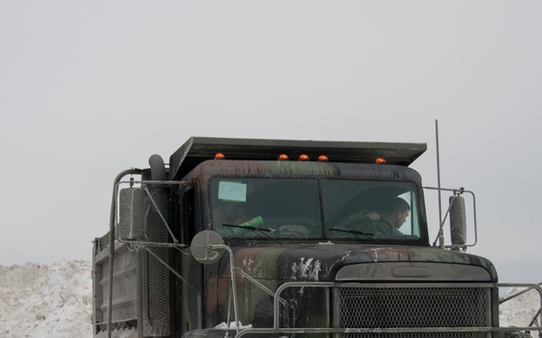 A large military-style dump truck painted in olive drab/dark green is operating in a snowy environment, surrounded by large piles of plowed snow. The truck appears to be actively engaged in snow removal or transport operations, with at least one occupant visible in the cab. The vehicle shows signs of heavy use with mud and snow accumulation on its exterior.