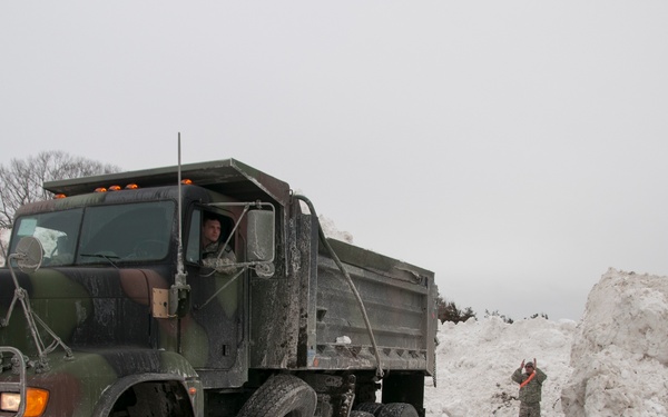 A military camouflage-painted dump truck is operating in a snow removal operation, surrounded by large piles of accumulated snow. One person is visible in the truck cab while another person in military-style clothing with an orange safety vest stands near a snow pile, appearing to direct the truck. The overcast sky and muddy, snow-covered ground suggest active winter storm recovery operations.