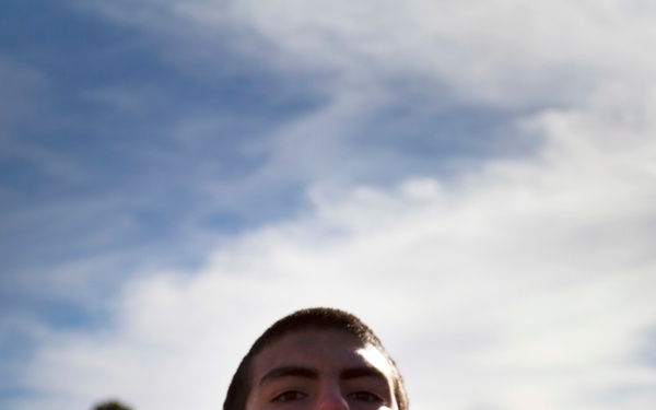 A young man is holding a dark brown book or trophy up to his chin/mouth level outdoors. The photo is taken from a low angle, emphasizing the sky and clouds in the background. A scoreboard structure is partially visible in the upper right corner.