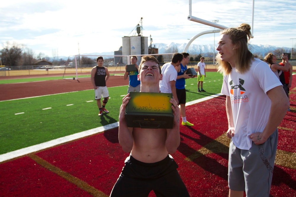A group of young athletes are gathered on a red artificial turf athletic field during what appears to be an outdoor practice or training session. A shirtless young man in the foreground is straining to lift or hold a yellow and black box, while a taller athlete in a white Under Armour shirt watches nearby. Snow-capped mountains and industrial structures are visible in the background.