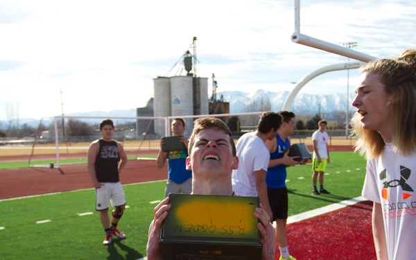 A group of young athletes are gathered on a red artificial turf athletic field during what appears to be an outdoor practice or training session. A shirtless young man in the foreground is straining to lift or hold a yellow and black box, while a taller athlete in a white Under Armour shirt watches nearby. Snow-capped mountains and industrial structures are visible in the background.