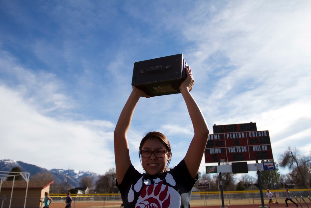 A young woman wearing glasses and a black t-shirt with a bear paw graphic is holding a dark rectangular box (likely a boombox or speaker) above her head and smiling. She is standing on or near an athletic track field with a football scoreboard visible in the background. Snow-capped mountains are visible in the distance under a partly cloudy blue sky.
