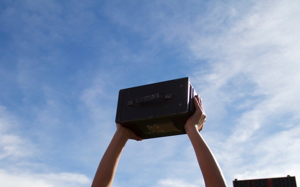 A young woman wearing glasses and a black t-shirt with a bear paw graphic is holding a dark rectangular box (likely a boombox or speaker) above her head and smiling. She is standing on or near an athletic track field with a football scoreboard visible in the background. Snow-capped mountains are visible in the distance under a partly cloudy blue sky.
