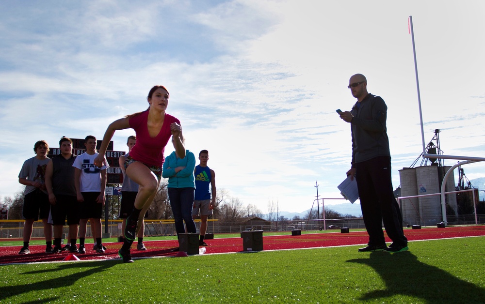 A female athlete is captured mid-sprint on a red track surface at what appears to be a high school or collegiate athletic facility. A coach or official stands nearby holding papers and a device, likely timing or recording the performance, while a group of athletes watches in the background. The setting suggests a track and field practice or tryout session.