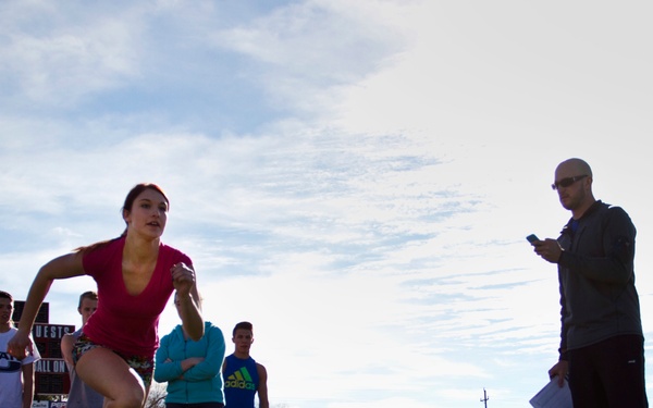A female athlete is captured mid-sprint on a red track surface at what appears to be a high school or collegiate athletic facility. A coach or official stands nearby holding papers and a device, likely timing or recording the performance, while a group of athletes watches in the background. The setting suggests a track and field practice or tryout session.