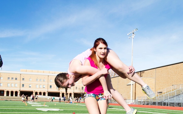 Two women are participating in what appears to be a wife-carrying or partner-carrying race on an artificial turf football field. One woman is carrying another on her back while running, as a male official or coach observes nearby holding a clipboard and phone. The event appears to be an organized athletic competition taking place at a school or university stadium.