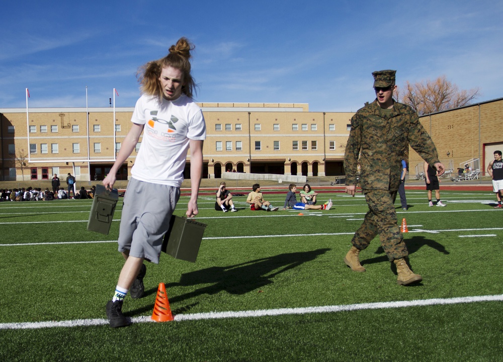 A young person with long red hair carries military-style ammunition cans across a football field while a U.S. Marine in full camouflage uniform walks alongside them. The activity appears to be a military fitness demonstration or recruiting event at a school, with other students visible in the background sitting and observing. The setting is an outdoor athletic field adjacent to a brick school building.