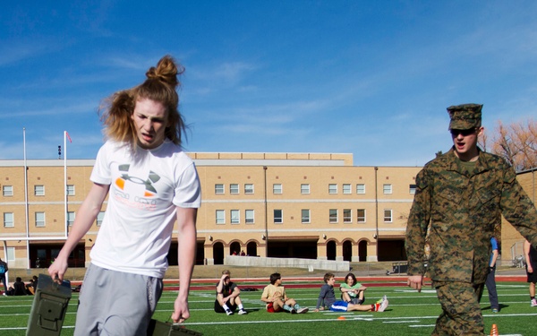 A young person with long red hair carries military-style ammunition cans across a football field while a U.S. Marine in full camouflage uniform walks alongside them. The activity appears to be a military fitness demonstration or recruiting event at a school, with other students visible in the background sitting and observing. The setting is an outdoor athletic field adjacent to a brick school building.