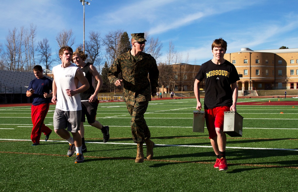 A group of young men are participating in what appears to be a military fitness recruitment or ROTC-style physical training event on an outdoor athletic field. A uniformed U.S. Marine in woodland camouflage (MARPAT) is leading or supervising the activity. One participant in a Missouri Adidas shirt is carrying two military-style ammunition cans as part of the exercise.