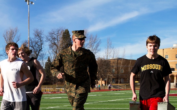 A group of young men are participating in what appears to be a military fitness recruitment or ROTC-style physical training event on an outdoor athletic field. A uniformed U.S. Marine in woodland camouflage (MARPAT) is leading or supervising the activity. One participant in a Missouri Adidas shirt is carrying two military-style ammunition cans as part of the exercise.