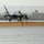 2015 Australian International Airshow and Aerospace & Defence Exposition A fifth-generation stealth fighter aircraft, consistent with an F-22 Raptor, is taxiing on a wet runway during rainy conditions. The aircraft is viewed head-on, with landing lights illuminated and water visible on the tarmac surface. An orange and yellow safety barrier or arresting gear net is visible in the foreground.