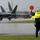 2015 Australian International Airshow and Aerospace & Defence Exposition A ground marshal wearing a high-visibility yellow 'Airshow Tarmac' vest is directing an F-22 Raptor stealth fighter jet on a wet tarmac during heavy rain. The marshal is holding two red signaling paddles with both arms raised. The scene appears to be at a civilian airshow, with a commercial aircraft visible in the background.