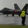 2015 Australian International Airshow and Aerospace & Defence Exposition A ground marshal wearing a high-visibility yellow vest labeled 'AIRSHOW TARMAC' is directing a dark gray stealth fighter jet — identifiable as an F-22 Raptor — on a wet tarmac during heavy rain. The marshal is holding orange signal wands in both hands to guide the aircraft. The scene appears to be at a military airshow, likely in Australia based on contextual clues.