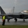 2015 Australian International Airshow and Aerospace & Defence Exposition An F-22 Raptor stealth fighter jet is taxiing on a wet runway during overcast, rainy conditions. A ground crew member wearing a high-visibility yellow vest and helmet stands on the tarmac, likely directing or monitoring the aircraft. The tail markings 'HH' and partial serial number '060' are clearly visible on the aircraft.
