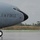 2015 Australian International Airshow and Aerospace & Defence Exposition A U.S. Air Force aircraft, marked with the number 1499, is taxiing on a wet tarmac under overcast skies. A ground crew member wearing a high-visibility yellow vest is directing the aircraft using orange marshalling wands. The setting appears to be a military or civilian airfield with fencing, trees, and utility poles visible in the background.
