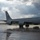 2015 Australian International Airshow and Aerospace & Defence Exposition A U.S. Air Force KC-135 Stratotanker aerial refueling aircraft is parked on a wet tarmac, likely following recent rainfall. The aircraft bears the tail code 'HH' and serial number '11499', with the 'U.S. AIR FORCE' inscription clearly visible on the fuselage. The dramatic overcast sky with rays of light breaking through clouds provides a striking backdrop to the scene.