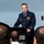 2015 Australian International Airshow and Aerospace & Defence Exposition A senior military officer in a dark navy dress uniform is delivering a speech at a podium during what appears to be an outdoor airshow event in 2015. The podium displays partial text reading 'NATIONAL AIRSHOW' and 'DEFENCE EXPOSITION' with 'ION 2015' visible. An audience is visible in the foreground with their backs to the camera.