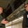 2015 Australian International Airshow and Aerospace & Defence Exposition Two men are engaged in conversation beneath what appears to be the open cargo bay or bomb bay of a large military aircraft. The man on the left is wearing an olive green flight suit with a squadron patch and a lanyard badge, while the man on the right is dressed in civilian attire with a blazer, dark shirt, sunglasses, and a visitor badge. The setting suggests a military aircraft tour or briefing event.