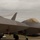 2015 Australian International Airshow and Aerospace & Defence Exposition Two F-22 Raptor stealth fighter jets are parked on a flight line at what appears to be an air show, with safety remove-before-flight tags still attached. In the background, two aircraft are performing a flying display, leaving white smoke trails against an overcast sky. The scene is consistent with a static display area at a military air show, with fencing, spectators, and support vehicles visible.