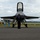 2015 Australian International Airshow and Aerospace & Defence Exposition A military jet fighter, consistent with an F-16 Fighting Falcon, is parked on a tarmac at what appears to be an airshow. Three ground crew personnel in dark uniforms are positioned around the aircraft — one directly behind it and one on each side — in a standard ground safety formation. Fire extinguishers and safety cones are visible, indicating active ground operations or pre/post-flight procedures.
