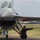 2015 Australian International Airshow and Aerospace & Defence Exposition Two ground crew members in black flight suits are marshaling an F-16 Fighting Falcon jet fighter on a tarmac. The crew member on the left has text on their back reading 'PACIFIC AIR FORCES DEMONSTRATION TEAM', indicating this is a demonstration or airshow event. A yellow ground support vehicle is visible in the background on the airfield.