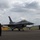 2015 Australian International Airshow and Aerospace & Defence Exposition Three ground crew members in matching dark jackets are saluting an F-16 Fighting Falcon jet fighter on a military airfield tarmac. A second F-16 is visible in the background to the right, along with what appears to be a larger propeller-driven aircraft further in the distance. The scene appears to be a formal send-off or arrival ceremony under an overcast sky.