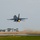 2015 Australian International Airshow and Aerospace & Defence Exposition A single-engine fighter jet, consistent with an F-16 Fighting Falcon, is captured moments after takeoff with afterburner fully engaged, producing a visible orange flame from the exhaust nozzle. The aircraft is in a steep climb attitude just above the runway surface, with jet exhaust blast disturbing water or wet debris on the runway behind it. The overcast sky and wet runway conditions suggest recent rainfall at the airfield.