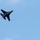 2015 Australian International Airshow and Aerospace & Defence Exposition A military jet fighter is captured in flight against a clear blue sky. The aircraft appears to be banking or performing a maneuver, showing its underside to the camera. The image is taken from a ground-level perspective looking upward.