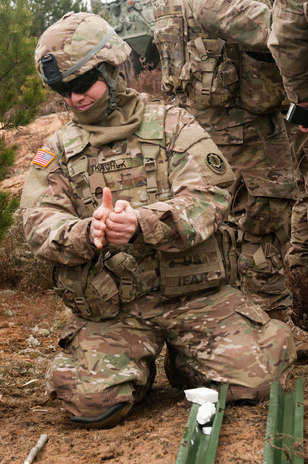 A U.S. Army soldier wearing MultiCam (OCP) camouflage uniform is kneeling on the ground in an outdoor wooded/sandy terrain environment. The soldier, identified by the name tape 'TKACHUK' on their chest, is gesturing with their hands while appearing to explain or demonstrate something. A second soldier in similar gear is partially visible in the background.