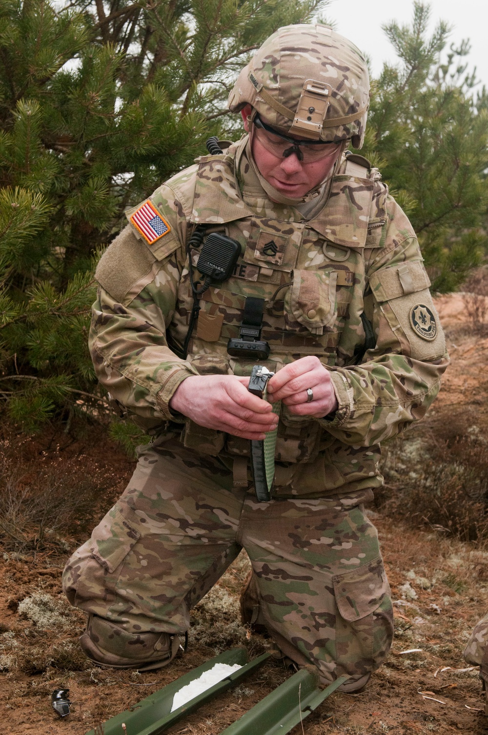 A U.S. Army soldier in MultiCam (OCP) camouflage uniform is kneeling on the ground in a wooded/scrubland environment, focused on manipulating a small object in his hands. He is wearing full combat gear including a helmet with a mounted device, body armor, and a radio. A green military item (possibly an antenna base or equipment component) is visible on the ground in front of him.