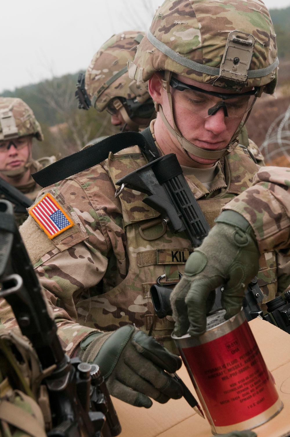 A U.S. Army soldier in MultiCam (OCP) camouflage uniform and combat helmet is handling a red cylindrical canister, likely a smoke grenade or similar training device. The soldier's name tape partially reads 'KIL-' and a U.S. flag patch is visible on the left shoulder. At least two other soldiers in similar gear are visible in the background.